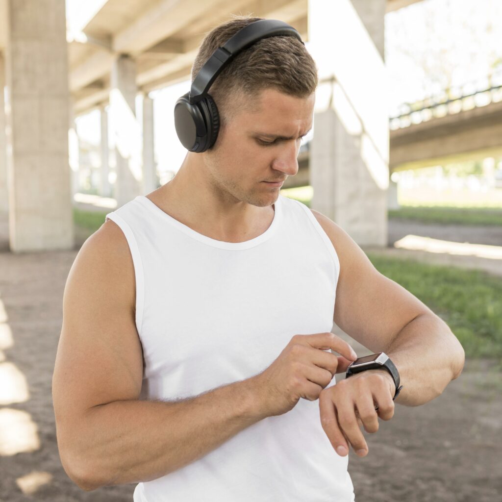 Homme avec un débardeur blanc et des écouteurs noirs, interagissant avec sa montre connectée avant de commencer une activité physique sous un pont.