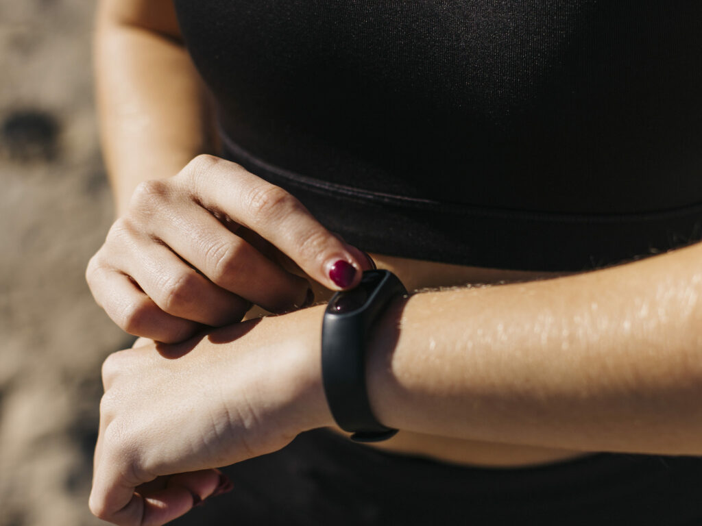 Femme sportive en tenue de fitness sur la plage, utilisant un bracelet connecté pour suivre son activité physique.