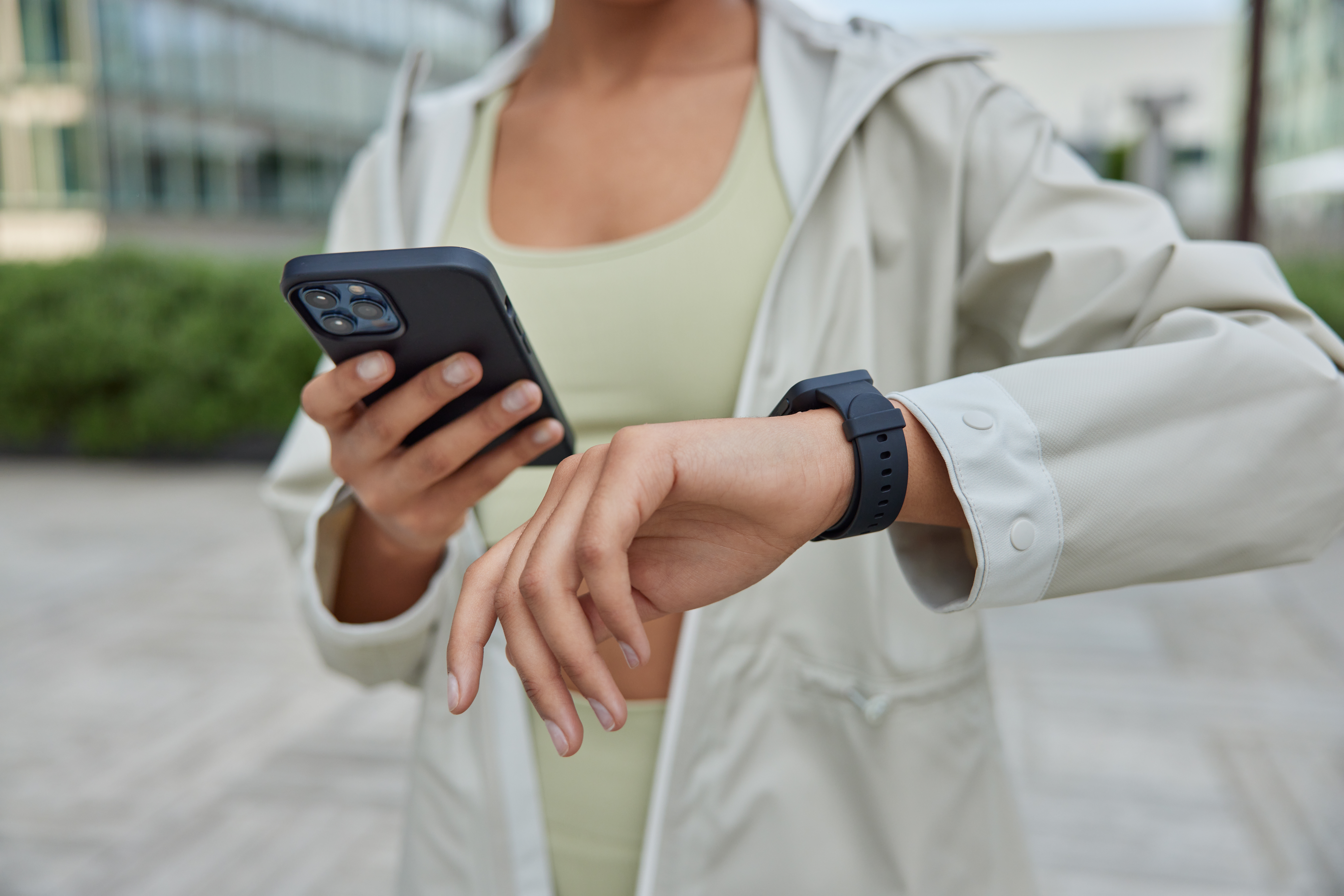 L'image montre une personne en tenue de sport, portant une veste légère beige et un bracelet connecté noir au poignet gauche. Elle tient un smartphone dans la main droite, semblant vérifier ou comparer des informations entre l'application mobile et le bracelet. Le décor en arrière-plan semble être un environnement urbain ou un parc avec des bâtiments modernes et des plantes, suggérant qu'elle est en extérieur, probablement avant ou après une activité physique.