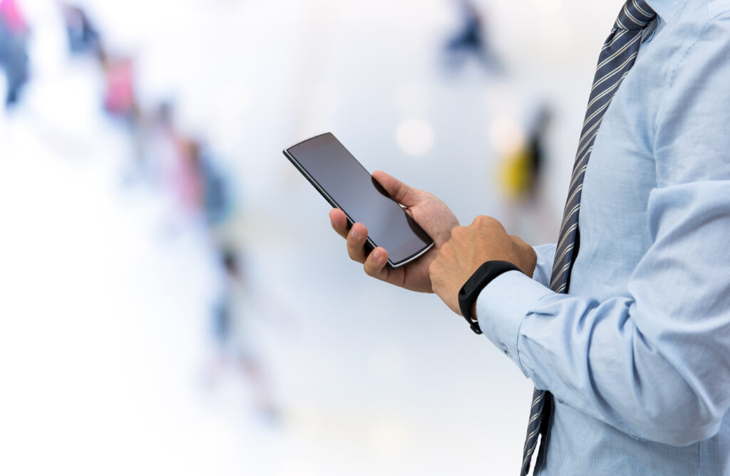 Cette image montre une personne en chemise bleue et cravate rayée, tenant un smartphone dans une main tout en portant un bracelet connecté noir à l'autre poignet. La personne interagit probablement avec l'écran du téléphone. L'arrière-plan est flou avec des formes de personnes suggérant un environnement public ou un espace de travail moderne, avec une forte luminosité ambiante. Le focus est mis sur l'usage simultané de la technologie portable (smartphone et bracelet connecté).