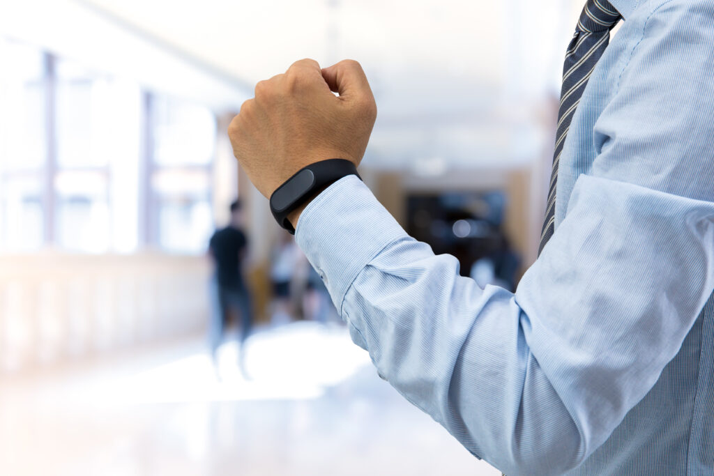 Cette image montre une personne portant une chemise bleue et une cravate à rayures, levant légèrement son poing pour montrer un bracelet connecté noir à son poignet. L'arrière-plan est flou, suggérant un environnement de bureau ou un espace intérieur lumineux avec des personnes indistinctes en arrière-plan. Le focus est mis sur le bracelet connecté et le bras, mettant en avant l'aspect technologique du wearable.